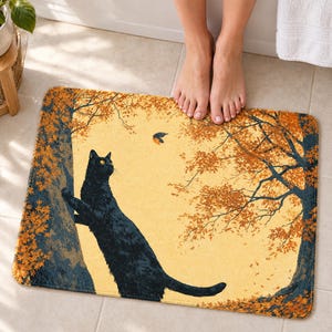 Climbing cat autumn bath mat on a tiled floor in front of a white bathtub: a cat reaches up a tree trunk toward a small bird between two trees with orange leaves; bare feet stand at the top edge nearest the tub.