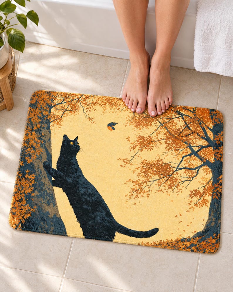 Climbing cat autumn bath mat on a tiled floor in front of a white bathtub: a cat reaches up a tree trunk toward a small bird between two trees with orange leaves; bare feet stand at the top edge nearest the tub.