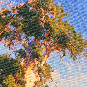 May include: A textured fabric print featuring a detailed landscape scene with a large tree displaying dense green and golden foliage against a bright blue sky. The surface shows a distinct woven canvas pattern with visible cross-hatch texture.