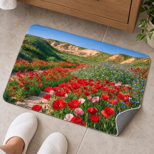 Poppy and wildflower bath mat on a gray tiled floor beside a wooden vanity, with feet in white slippers at the front edge; one corner flips up to show the gray anti-slip back.