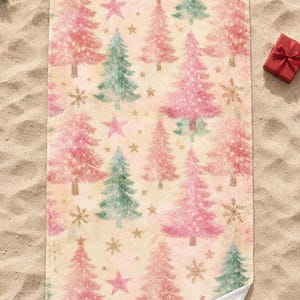 Pink Christmas tree beach towel laid flat on sand with sunglasses, a pine sprig and a small red gift box, one corner folded to show the white back.