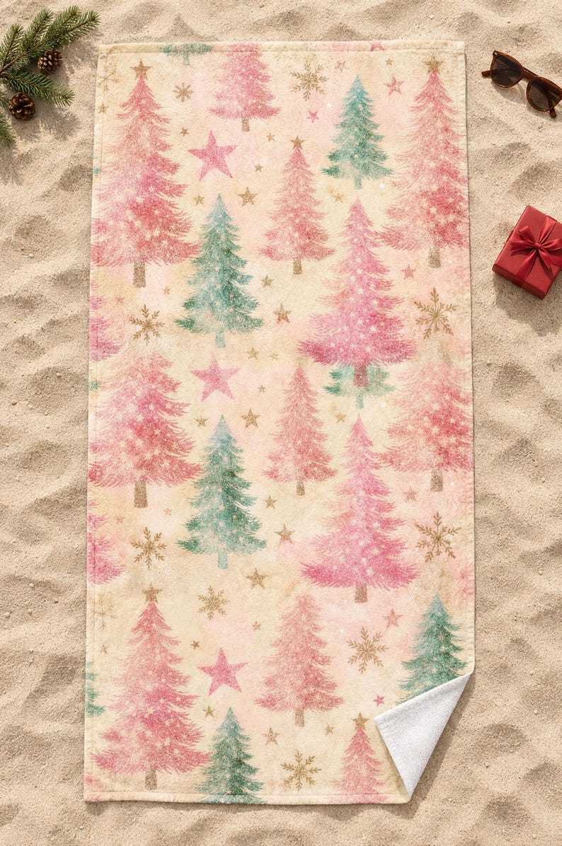 Pink Christmas tree beach towel laid flat on sand with sunglasses, a pine sprig and a small red gift box, one corner folded to show the white back.