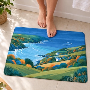 Cliffside cottage bath mat on a tiled floor in front of a white bathtub: a blue bay curves past green headlands and two white cottages among orange and blue shrubs; bare feet stand on the top edge nearest the tub.