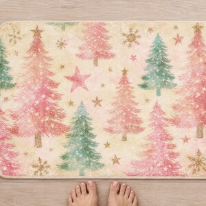 Pink Christmas tree bath mat seen from above on a light wood floor, with rolled white towels, a candle, a pine sprig and bare feet at the bottom edge.