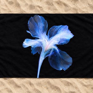以下が含まれることがあります： 中央に鮮やかな青と白のアイリスの花のグラフィックが描かれた、長方形の黒いビーチタオル。花びらは半透明で流れるような質感をしており、深い青の色合いが明るい白の中心部へと変化しています。