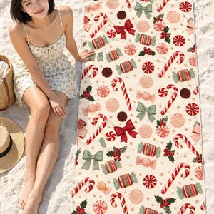 Candy cane Christmas beach towel laid on white sand, printed with candy canes, peppermint swirls, wrapped candies, bows and holly on cream, as a woman in a floral sundress sits beside it.
