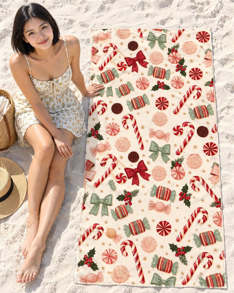 Candy cane Christmas beach towel laid on white sand, printed with candy canes, peppermint swirls, wrapped candies, bows and holly on cream, as a woman in a floral sundress sits beside it.