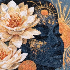 May include: A tufted textile wall hanging featuring a dark blue profile of a face adorned with large, textured cream and orange lotus flowers. The design includes a radiant sun motif in shades of orange and gold, with intricate sunburst patterns.