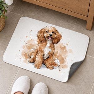 White bath mat with a sitting apricot Cavapoo puppy on a tiled floor by a wooden vanity; feet in white slippers rest on the tile in front and one corner flips up to show the gray anti-slip back.