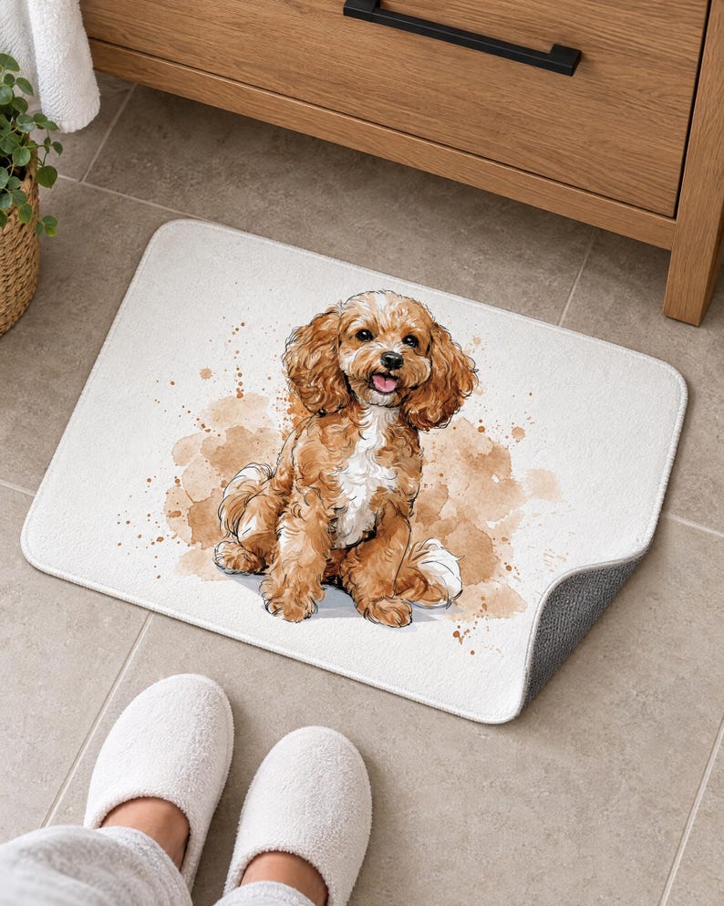 White bath mat with a sitting apricot Cavapoo puppy on a tiled floor by a wooden vanity; feet in white slippers rest on the tile in front and one corner flips up to show the gray anti-slip back.