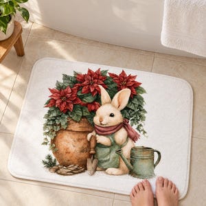 Watercolor bath mat with a cream bunny in a sage apron and scarf beside a pot of red poinsettias and a watering can on white, on a tiled floor by a tub with bare feet at the edge.
