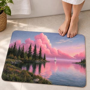 Pink cloud lake bath mat on a tiled floor in front of a white bathtub: pines on a grassy shore, a white sailboat and pink clouds reflected in calm water; bare feet stand on the top edge nearest the tub.
