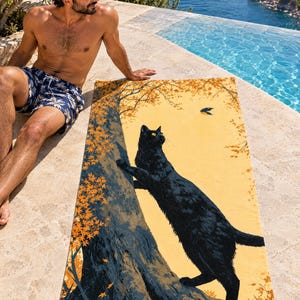 Fall cat beach towel on a stone pool edge above the sea, printed with a black cat climbing an autumn tree toward a small bird on gold, as a man in plaid swim shorts sits beside it.