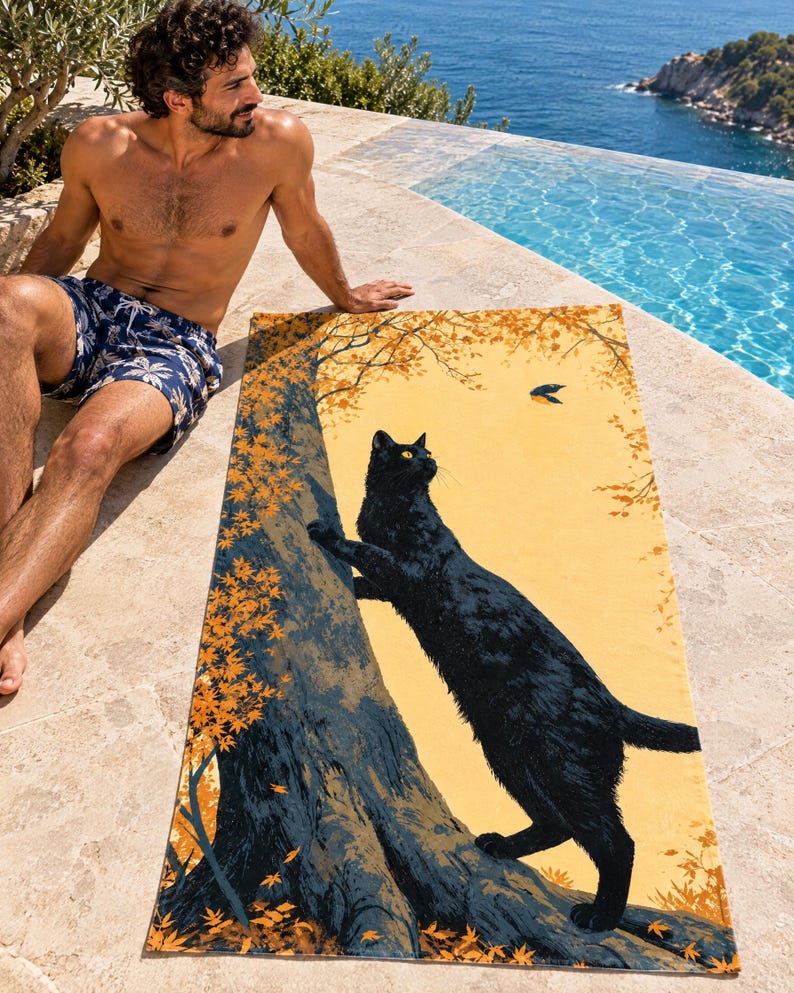 Fall cat beach towel on a stone pool edge above the sea, printed with a black cat climbing an autumn tree toward a small bird on gold, as a man in plaid swim shorts sits beside it.