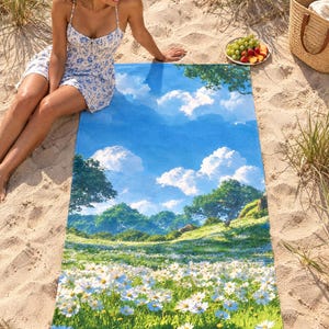 Daisy meadow beach towel laid on sand among dune grass, printed with white daisies and green hills under a blue sky with puffy clouds, as a woman in a floral sundress sits beside it.