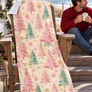Pink Christmas tree beach towel draped over a wooden deck chair by the ocean, with a smiling man in a red sweater holding a mug beside it.