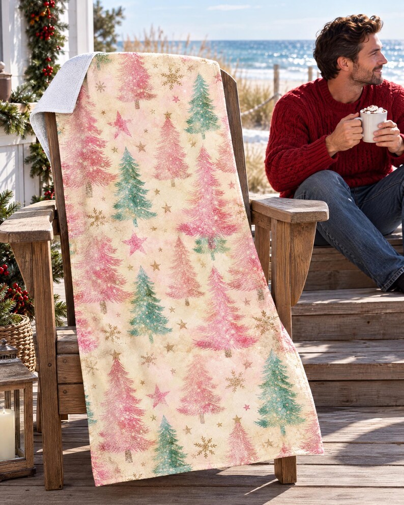 Pink Christmas tree beach towel draped over a wooden deck chair by the ocean, with a smiling man in a red sweater holding a mug beside it.