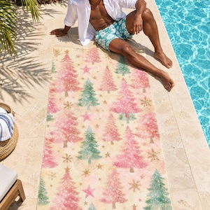 Pink Christmas tree beach towel laid on a sunny pool deck, printed with pink and sage trees, stars and snowflakes on cream, as a man in an open white shirt and swim shorts relaxes beside it.