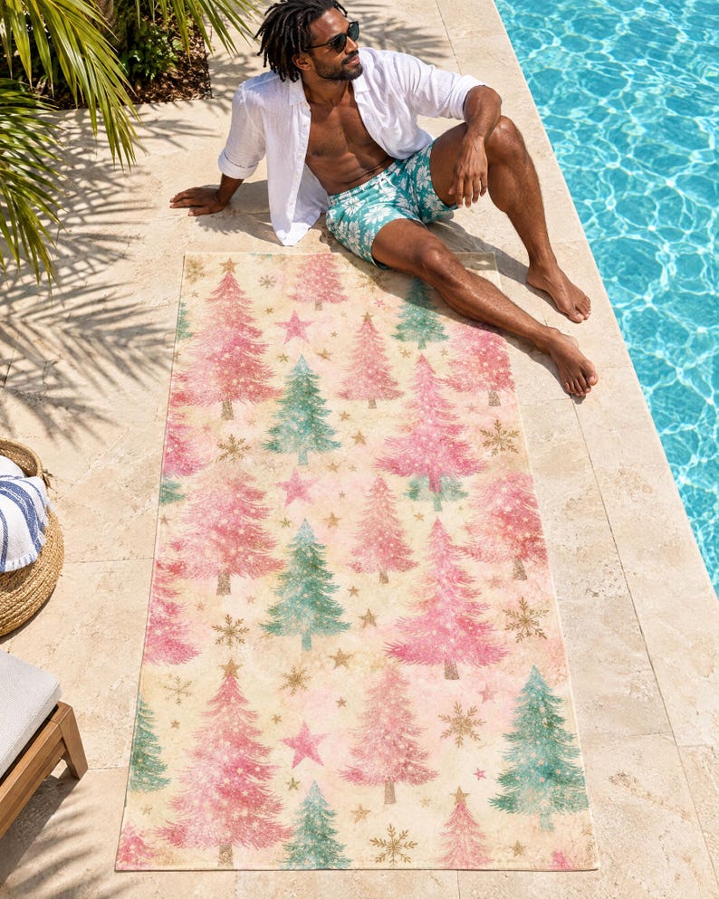 Pink Christmas tree beach towel laid on a sunny pool deck, printed with pink and sage trees, stars and snowflakes on cream, as a man in an open white shirt and swim shorts relaxes beside it.