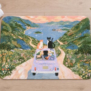 Overhead view of the whole cats road trip bath mat on a light wood floor, with rolled white towels, a lit candle and a small glass vase of wildflowers beside it.