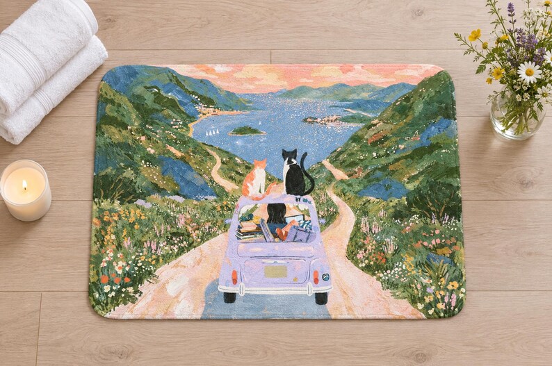 Overhead view of the whole cats road trip bath mat on a light wood floor, with rolled white towels, a lit candle and a small glass vase of wildflowers beside it.