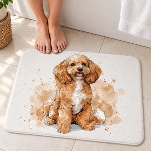 Cavapoo puppy bath mat on a tiled floor in front of a white bathtub: a curly apricot puppy with a white chest sits smiling among tan paint splashes on a white background; bare feet stand on the tile just above the mat.