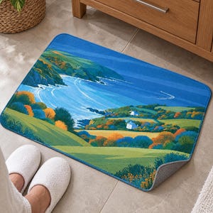 Cliffside cottage bath mat with a blue bay and green hills on a tiled floor by a wooden vanity; feet in white slippers rest on the tile in front and one corner flips up to show the gray anti-slip back.
