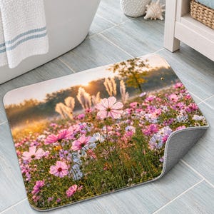 Peut inclure: Un tapis de bain rectangulaire orn&eacute; d'une impression photographique repr&eacute;sentant une prairie ensoleill&eacute;e remplie de fleurs de cosmos roses et de hautes herbes. L'image capture une lueur dor&eacute;e de lever de soleil sur le champ.