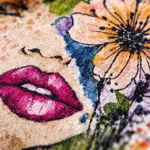 May include: A textile surface featuring a detailed embroidered illustration of a face with vibrant magenta lips, a large peach and yellow flower with black outlines, and green foliage accents against a textured beige background.