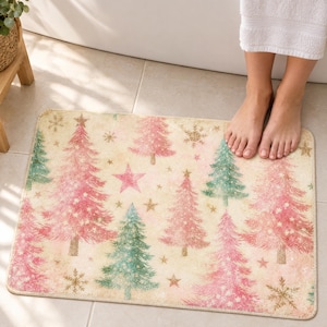 Pink Christmas tree bath mat with pink and sage trees, stars and gold snowflakes on cream, on a tiled bathroom floor by a tub with bare feet resting on it.
