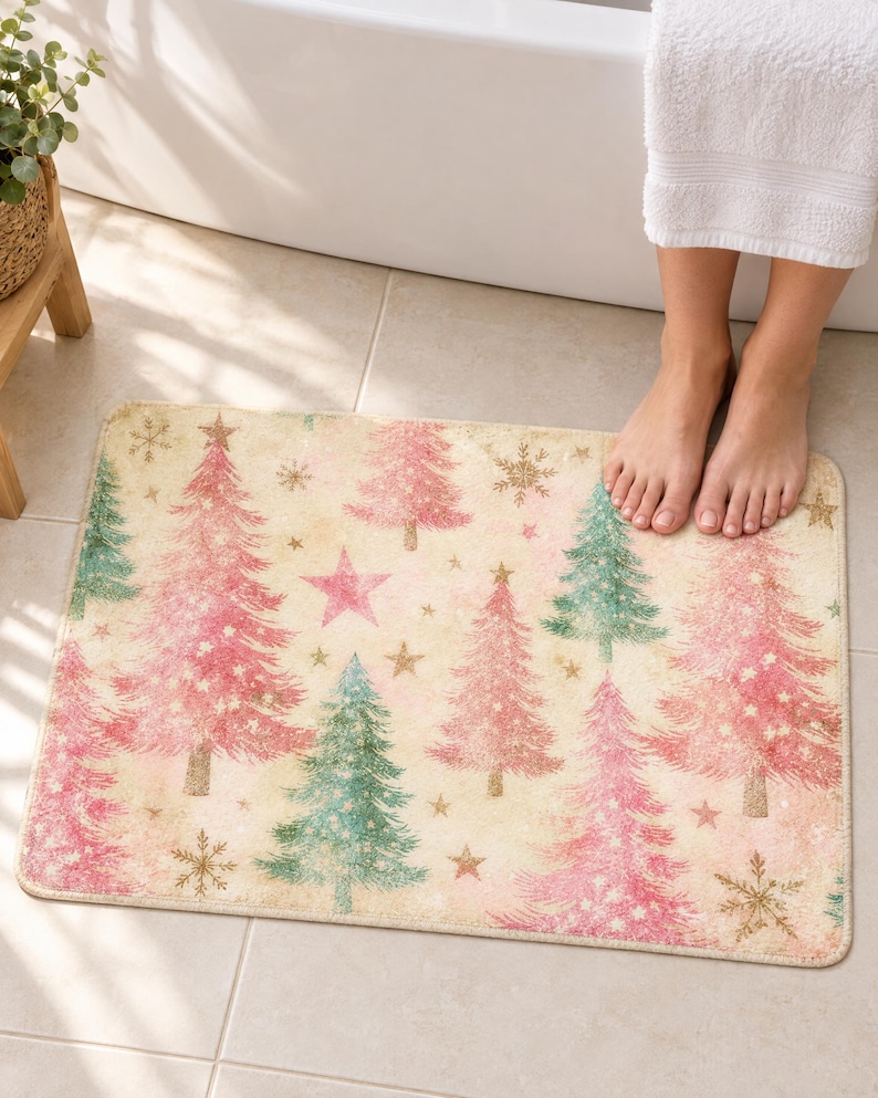 Pink Christmas tree bath mat with pink and sage trees, stars and gold snowflakes on cream, on a tiled bathroom floor by a tub with bare feet resting on it.