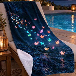 May include: A rectangular beach towel featuring a dark blue night sky and ocean scene. The design displays numerous glowing, multi-coloured origami cranes floating upward from the water towards a starry expanse, with a white underside visible.
