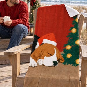 Sleeping beagle Santa hat beach towel draped over a wooden deck chair by the ocean, with a smiling man in a red sweater holding a mug nearby.