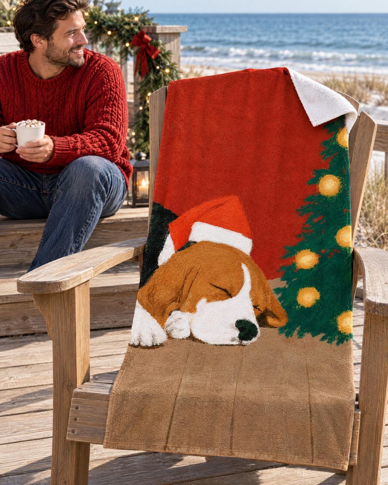 Sleeping beagle Santa hat beach towel draped over a wooden deck chair by the ocean, with a smiling man in a red sweater holding a mug nearby.