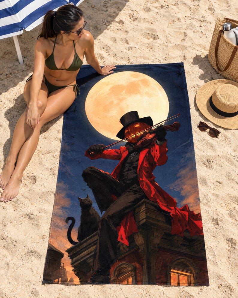 Halloween pumpkin violinist beach towel laid on sand, printed with a pumpkin-headed figure in a red coat and top hat playing a violin before a full moon, as a woman in a bikini sits beside it.