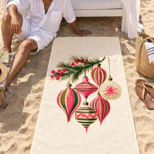 Retro Christmas ornament beach towel laid on sand by a cabana, printed with pink, red and olive glass ornaments hanging from a pine sprig on cream, as a man in white linen sits beside it.