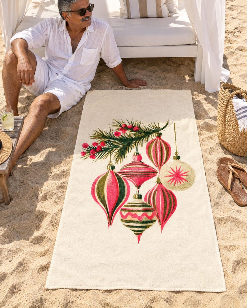 Retro Christmas ornament beach towel laid on sand by a cabana, printed with pink, red and olive glass ornaments hanging from a pine sprig on cream, as a man in white linen sits beside it.