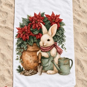 Poinsettia bunny beach towel laid flat on sand with sunglasses, a pine sprig and a small red gift box, one corner folded to show the white back.