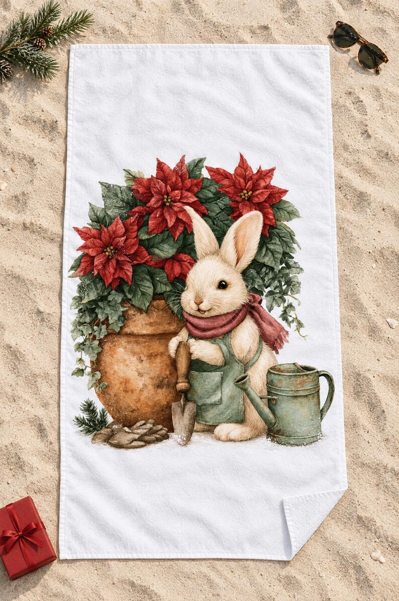 Poinsettia bunny beach towel laid flat on sand with sunglasses, a pine sprig and a small red gift box, one corner folded to show the white back.