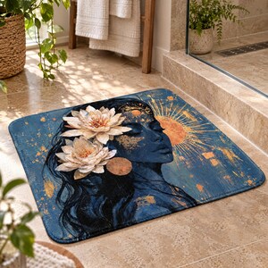 May include: A rectangular bath mat featuring a dark blue background with a profile illustration of a person adorned with two large white lotus flowers in their hair, accented by golden sun motifs and scattered gold leaf textures across the surface.