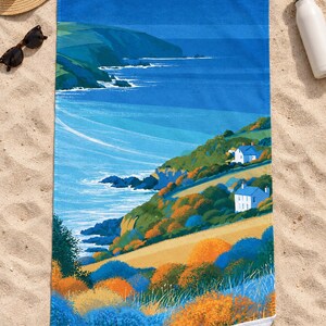 Top-down view of the whole sea cliff cottage beach towel laid flat on sand, with a plain deep blue band across the top above the headland, a straw hat, sunglasses and a white water bottle; one corner folds back to the white back.