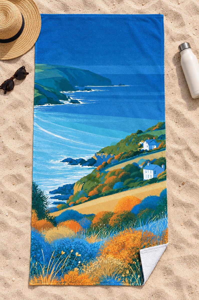 Top-down view of the whole sea cliff cottage beach towel laid flat on sand, with a plain deep blue band across the top above the headland, a straw hat, sunglasses and a white water bottle; one corner folds back to the white back.