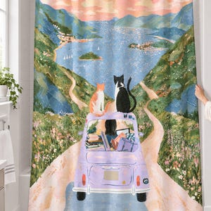 Lavender car cats shower curtain over a bathtub in a white-tiled bathroom: an orange cat and a black and white cat sit on a lavender car driven by a dark-haired driver down a road to a blue bay; a hand pulls back the right edge.