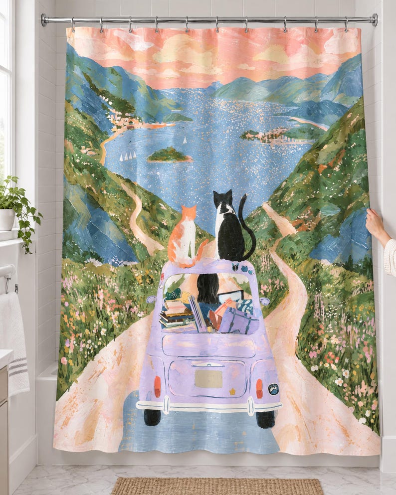 Lavender car cats shower curtain over a bathtub in a white-tiled bathroom: an orange cat and a black and white cat sit on a lavender car driven by a dark-haired driver down a road to a blue bay; a hand pulls back the right edge.