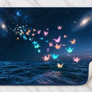 May include: A rectangular bath mat featuring a dark blue night sky filled with stars and galaxies above a dark ocean. A flock of glowing, multi-coloured origami cranes in shades of pink, orange, and blue flies across the scene.