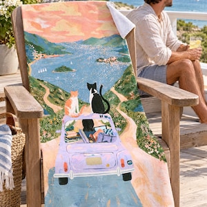 Two cats vintage car beach towel, the dark-haired driver visible in the lavender car, draped over a wooden Adirondack chair on a seaside deck with a corner folded to the white back; a man in sunglasses sits nearby with a drink.