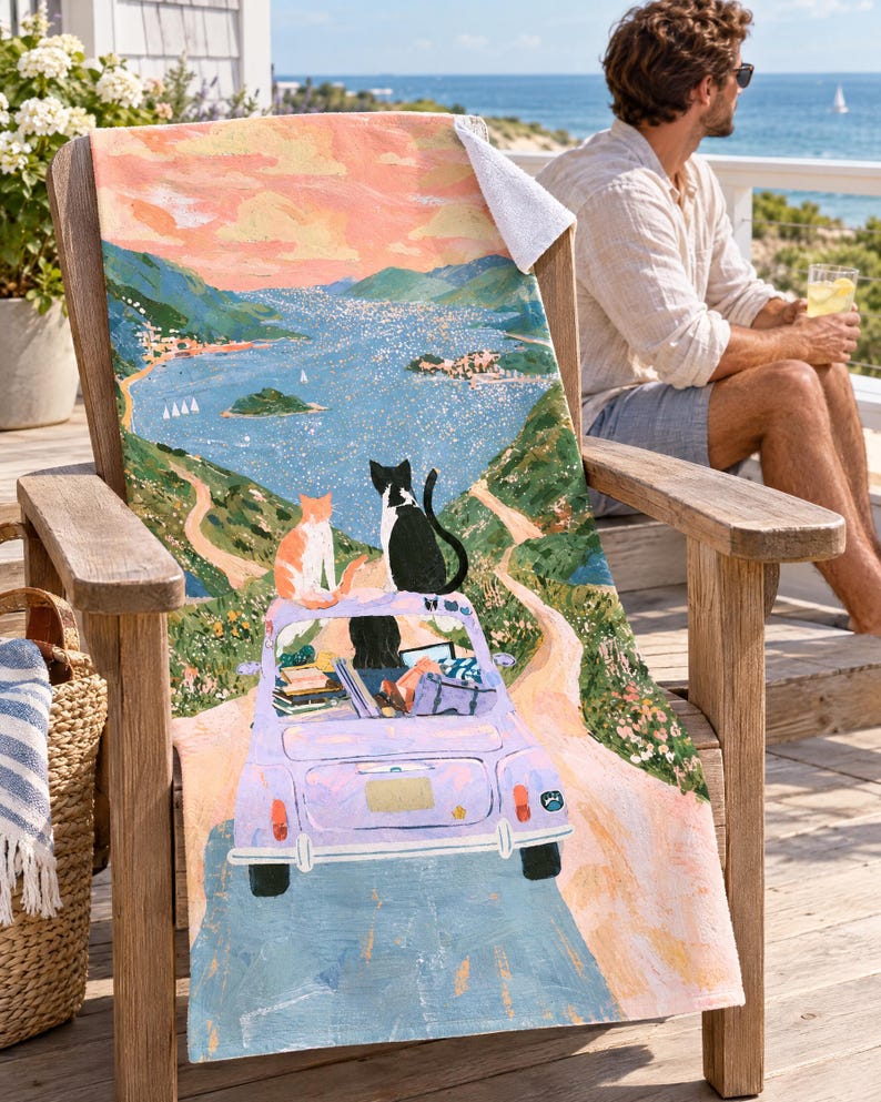 Two cats vintage car beach towel, the dark-haired driver visible in the lavender car, draped over a wooden Adirondack chair on a seaside deck with a corner folded to the white back; a man in sunglasses sits nearby with a drink.