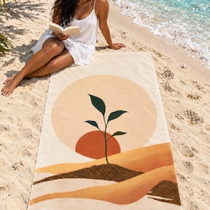 Boho sun beach towel laid on sand by the water, printed with a peach sun, a small green sprout and layered mustard dunes, as a woman in a white sundress and sun hat reads a book beside it.