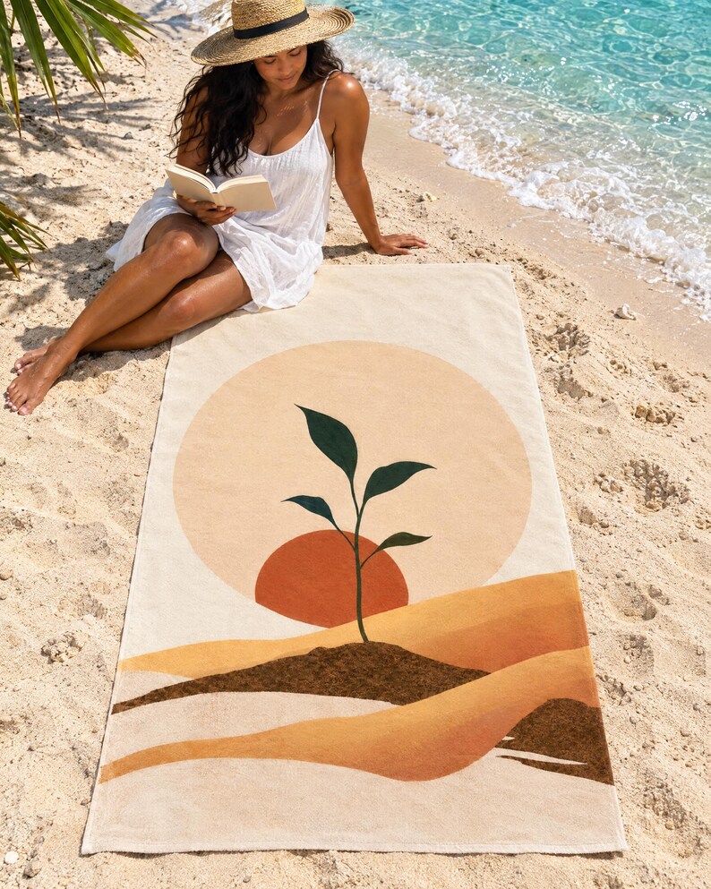 Boho sun beach towel laid on sand by the water, printed with a peach sun, a small green sprout and layered mustard dunes, as a woman in a white sundress and sun hat reads a book beside it.