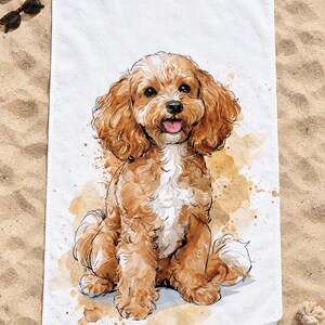 Top-down view of the whole watercolor Cavapoo beach towel laid flat on sand, the seated puppy surrounded by plain white space, with a straw hat, sunglasses and a rope toy; one corner folds back to the white back.
